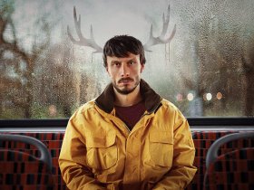 Donny sitting on the bus wearing a yellow jacket, reindeer ears in the window in a scene from Baby Reindeer.