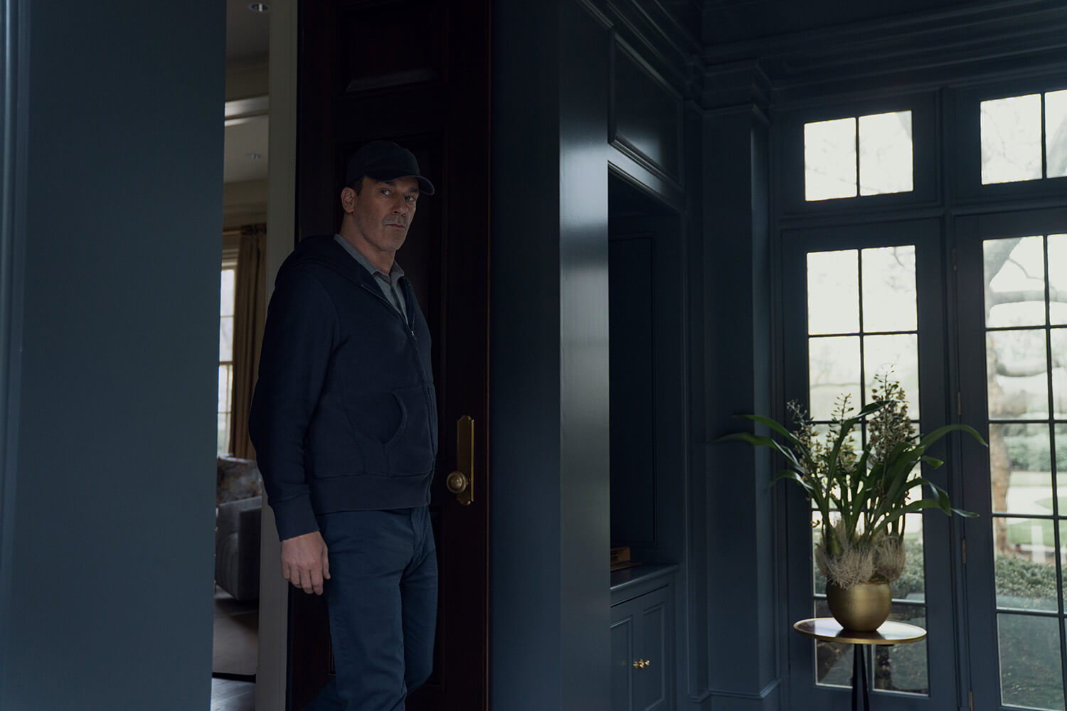 Coop standing by a door with a baseball hat looking suspicious in Your Friends and Neighbors.