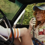 Charlie sitting in her car, leg up talking on a radio in Poker Face.