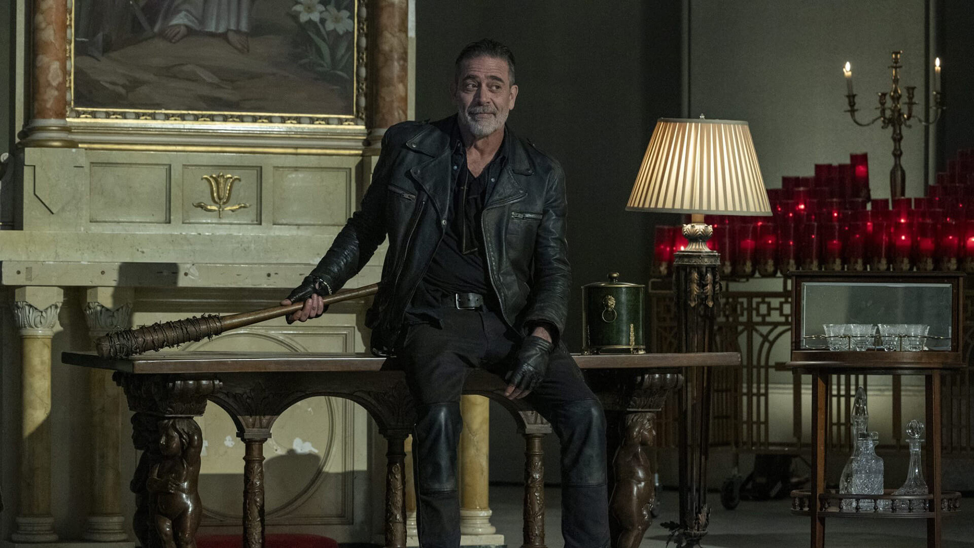 Negan sitting on a table with a barbed wire bat in The Walking Dead: Dead City.