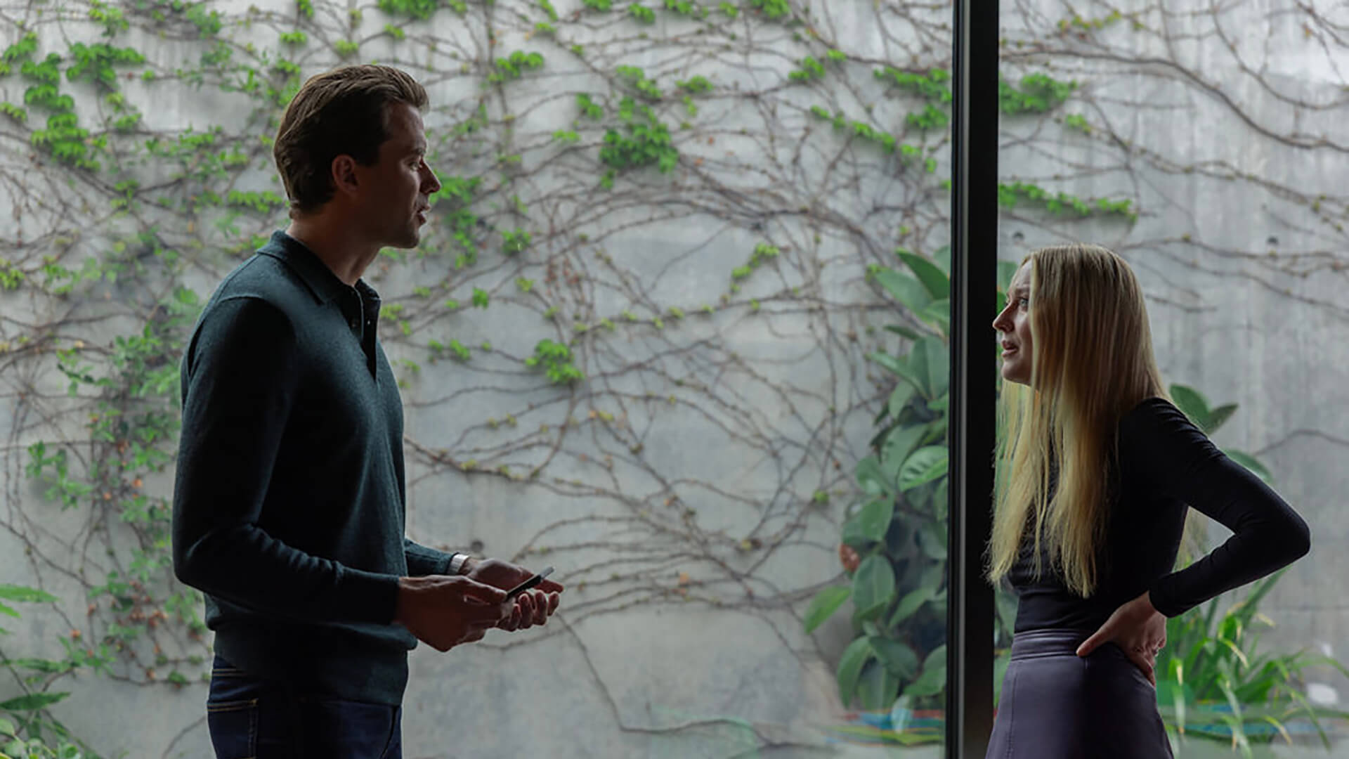 A man and woman talking by a large window in All Her Fault.