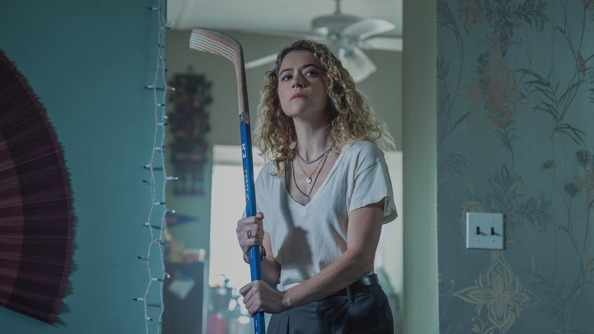 Paula standing at a doorway holding a hockey stick in Maximum Pleasure Guaranteed.