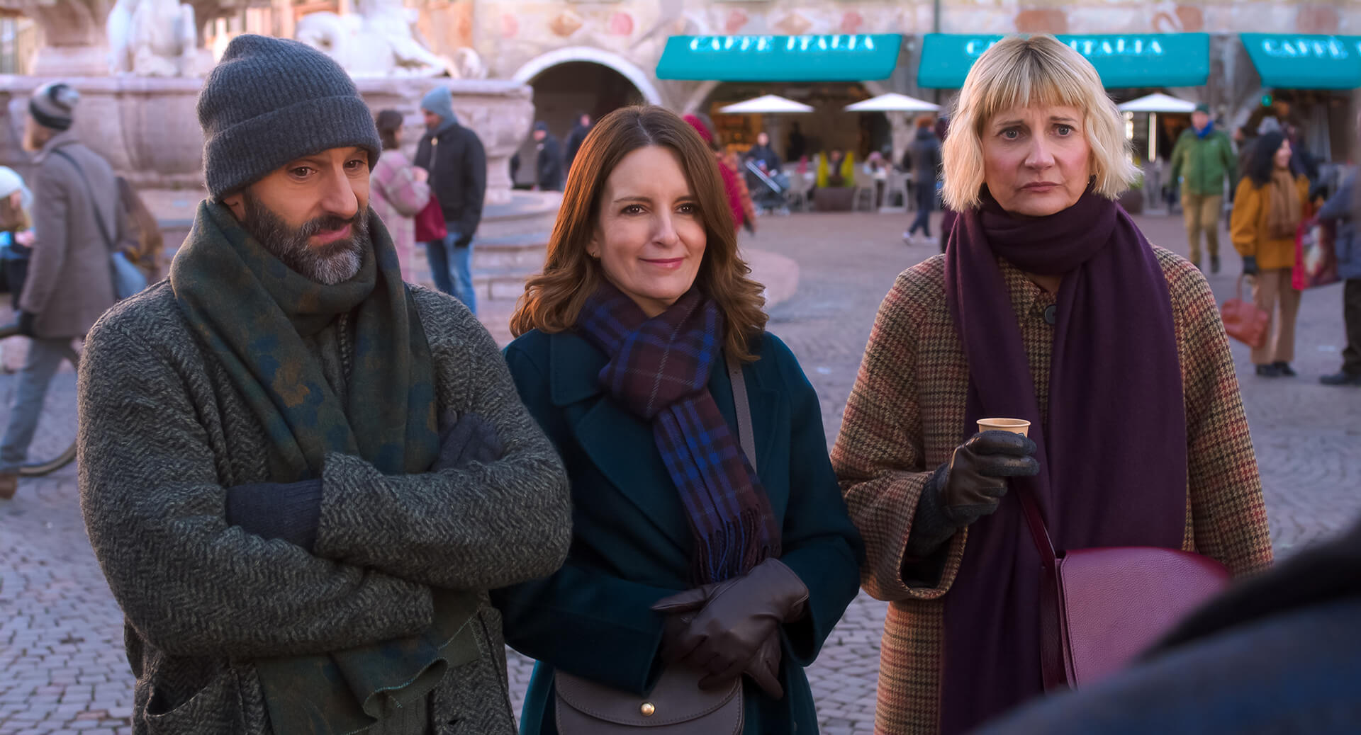 "Three adults in winter clothing stand together in a European outdoor plaza, with a cafe and people in the background. They appear to be having a casual conversation on a chilly day."