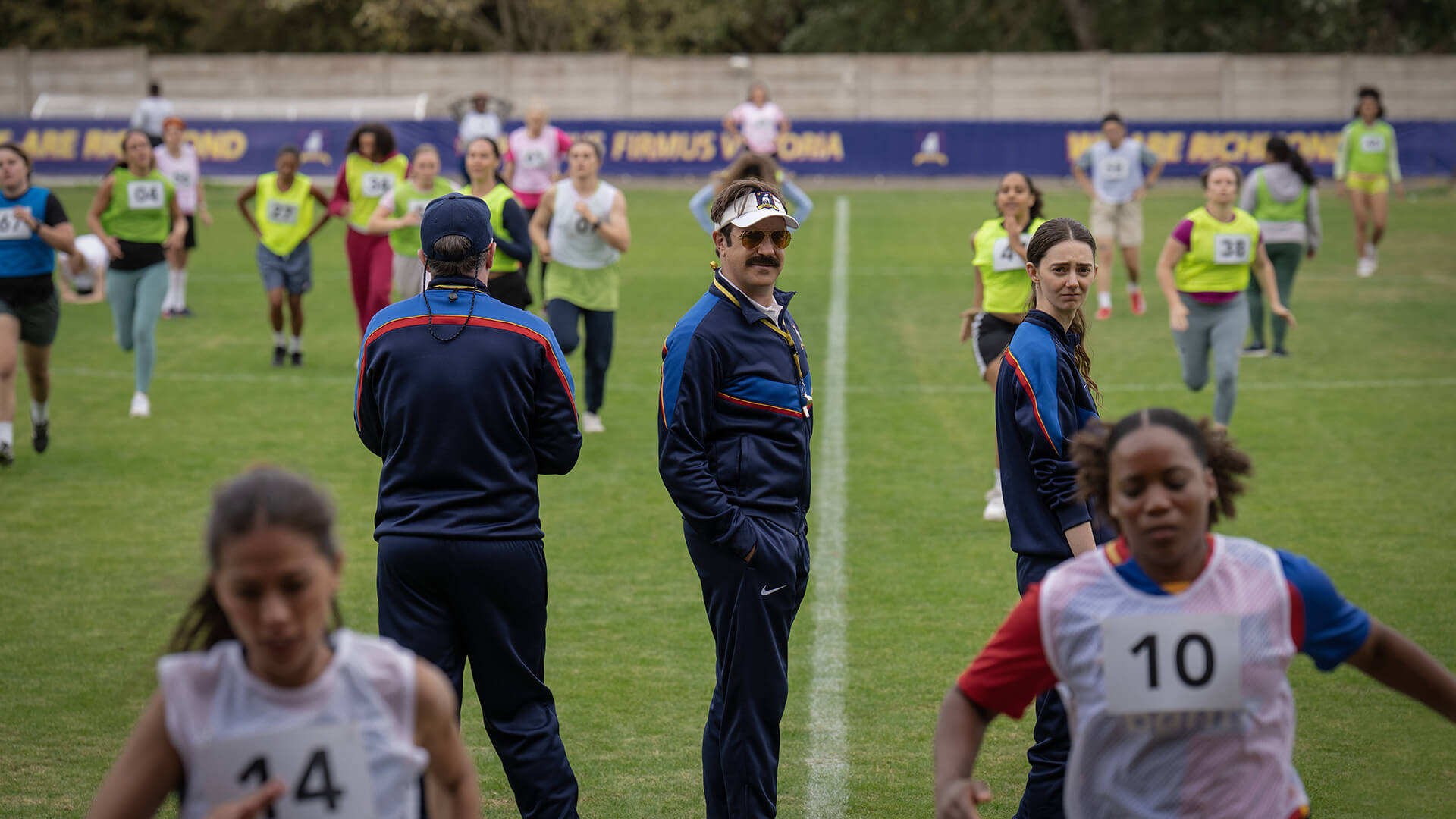 Ted on the field with the other coaches as the women run drills in Ted Lasso.