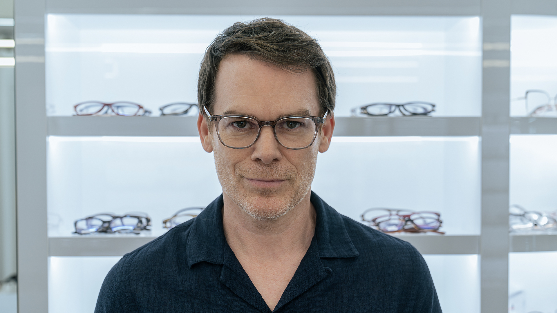 Dexter smiling while trying on glasses in a shop in Dexter: Resurrection.