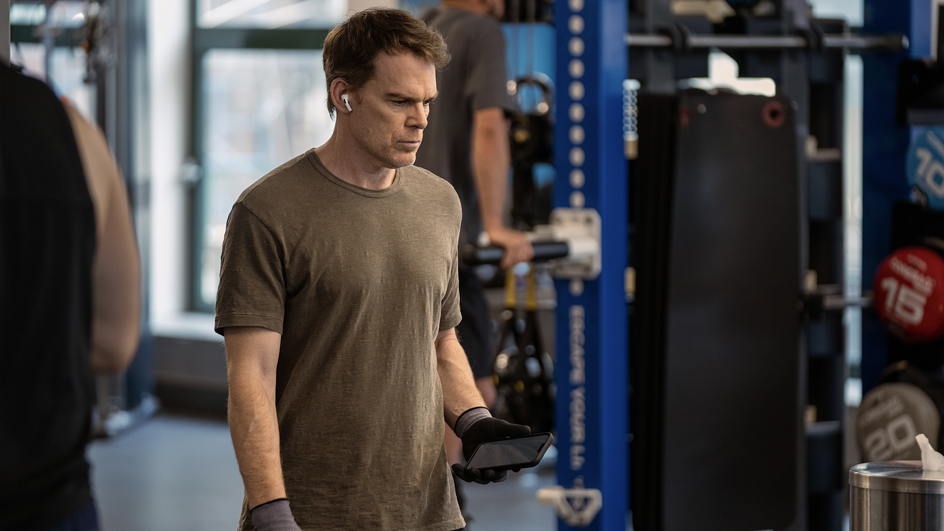 Dexter at the gym wearing earbuds and holding his phone in Dexter: Resurrection.