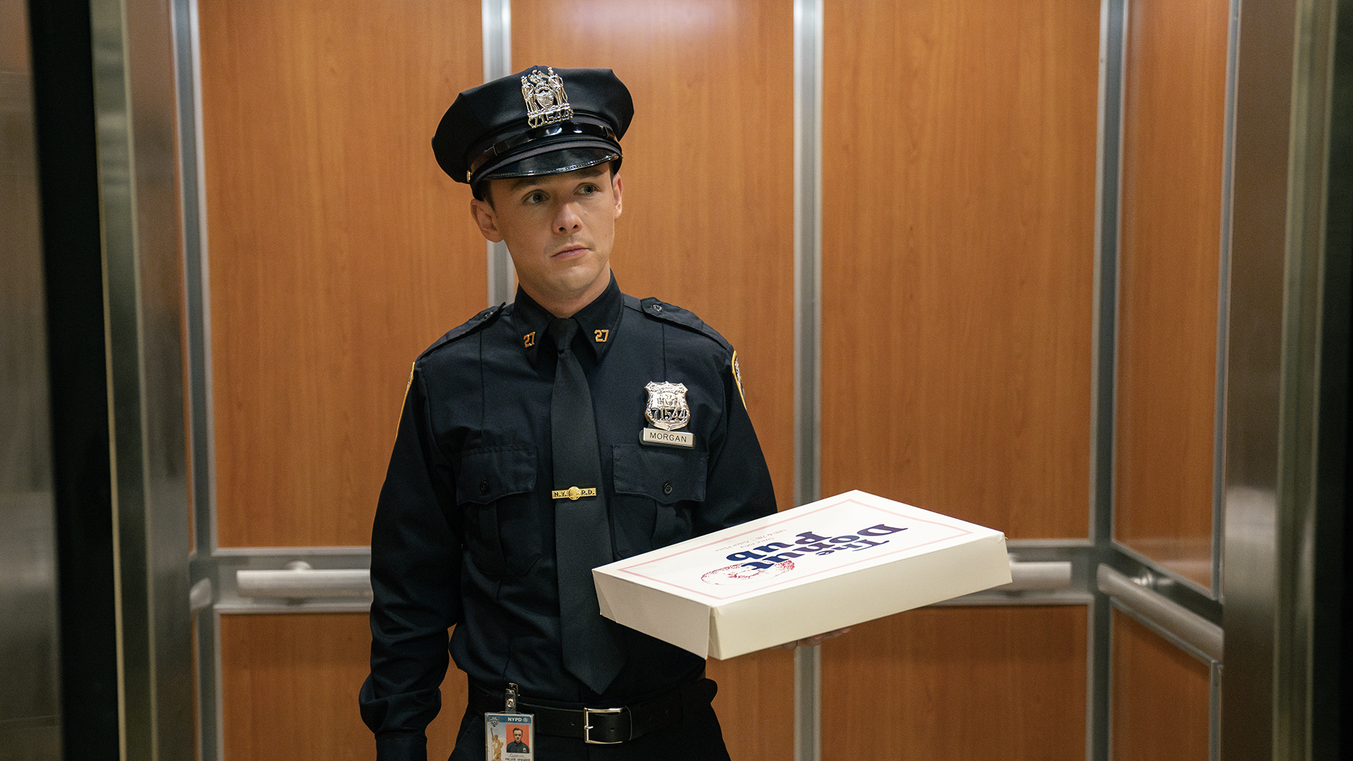 Harrison in a police uniform holding a box of donuts in the elevator in Dexter: Resurrection.
